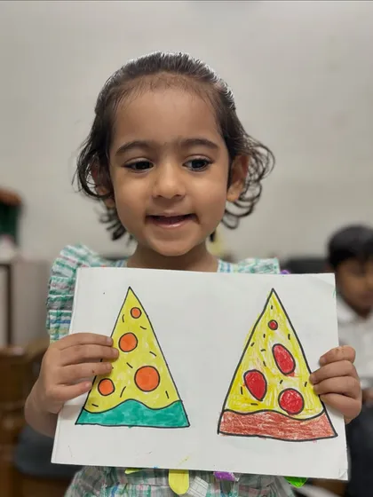 Learning shapes can be delicious. A junior KG student happily shows her drawing of pizza slices, a fun exercise we do to practice triangles and circles while learning to colour within the lines.