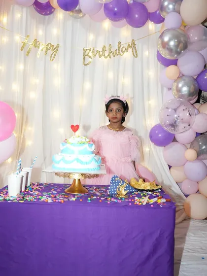 A happy birthday girl enjoying her special day. This cozy home setup features a backdrop of white curtains and warm fairy lights, framed by a balloon arch in pastel pink, purple, and silver.