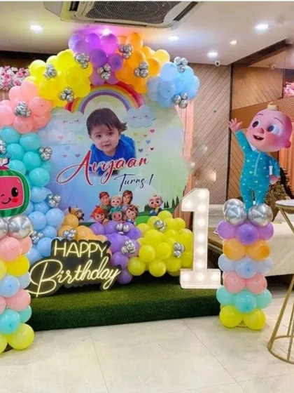 A bright and cheerful Cocomelon theme for a first birthday party. This setup includes a custom backdrop with the birthday boy's photo, colorful balloon arrangements, character cutouts, a 'Happy Birthday' neon sign, and a light-up number '1'.