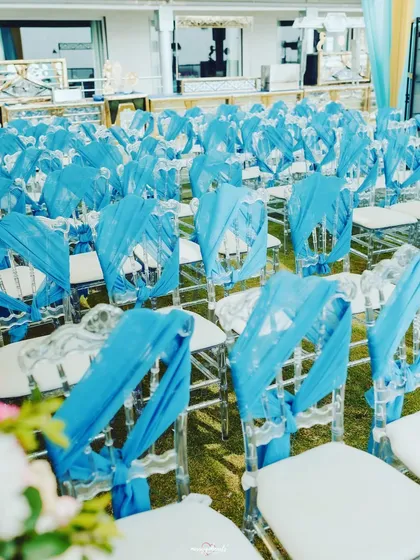 Details of a Haldi seating arrangement. The clear Chiavari chairs are elegantly draped with bright blue fabric, adding a pop of color to the outdoor setup.