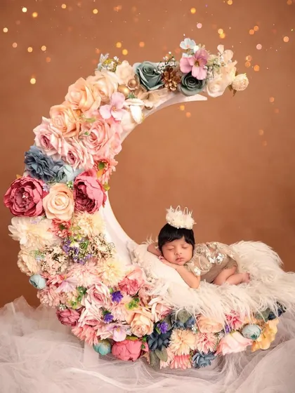 A newborn baby wearing a tiny crown sleeps on a crescent moon prop lavishly decorated with pastel-colored flowers. This is a perfect example of a high-concept, fantasy-themed newborn shoot.