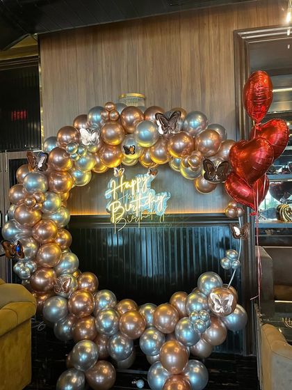 An elegant birthday backdrop with a butterfly theme. This circular arch is made with rose gold and silver balloons, accented with butterfly cutouts and a 'Happy Birthday' neon sign.