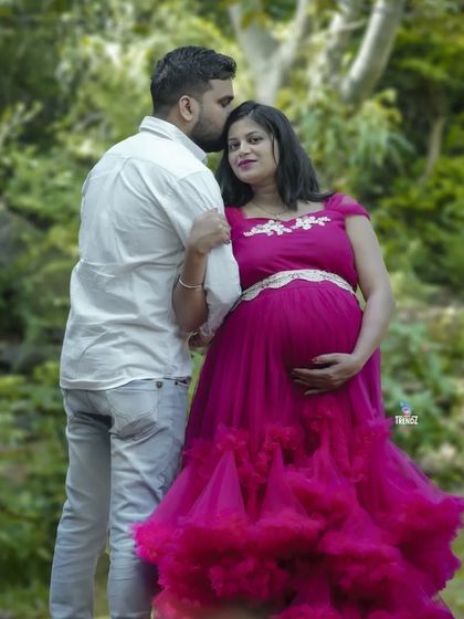 A tender moment during an outdoor maternity shoot. The partner's gentle kiss on the forehead shows a deep connection, complemented by the beautiful ruffled magenta gown.