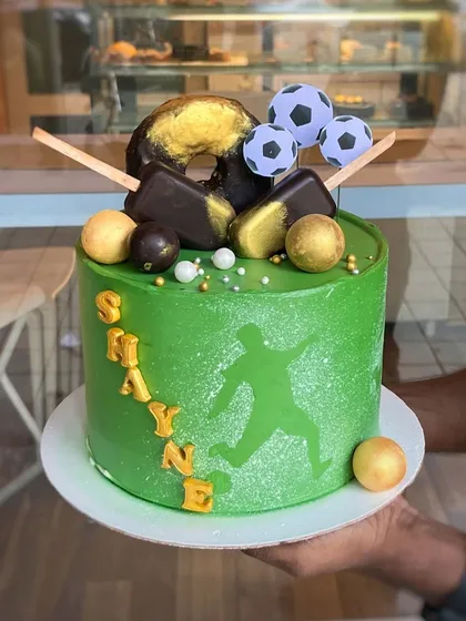 A football-themed cake for Shayne. The vibrant green cake features a silhouette of a player and is topped with chocolate-covered cakesicles, donuts, and fondant footballs for a fun and sporty design.