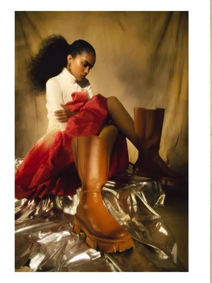 A creative shot focusing on the chunky leather boots, with the model seated on a silver-draped platform. This highlights key fashion details within the character study.