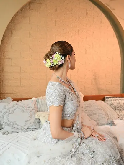 A beautiful shot of the bride seated, highlighting the elegant floral bun and the delicate details of her silver lehenga. The makeup is soft and luminous.