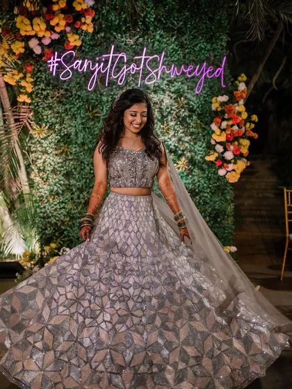 A candid moment of pure joy. This bride looks stunning at her Sangeet, posing in front of a custom neon sign in her shimmering silver lehenga.