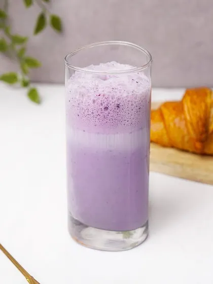 A layered blueberry smoothie in a glass, set against a clean white background with a croissant. This image effectively showcases a specialty beverage for a cafe or health food brand.
