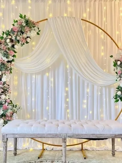 A classic and elegant engagement backdrop featuring a golden circular arch adorned with pastel pink and white flowers. The flowing white drapes and warm fairy lights create a soft, romantic ambiance, complemented by a plush tufted bench for seating.