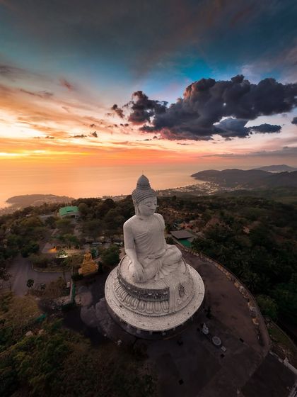 A wider drone shot of the Big Buddha, showing its majestic presence overlooking the coastline of Phuket at sunset.