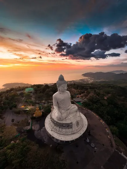A stunning aerial panorama of the Big Buddha complex in Phuket, with a beautiful sunset over the sea in the background.