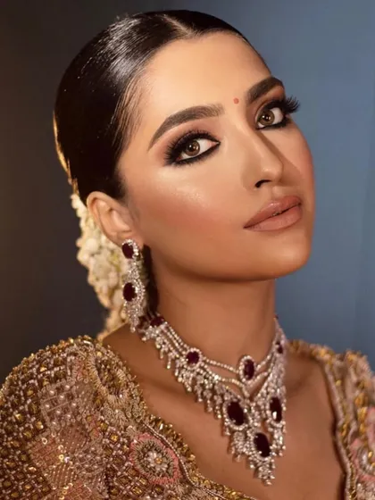 A close-up of a regal reception look. The makeup features a warm, brown smokey eye and a defined brow, paired with a beautiful ruby necklace.