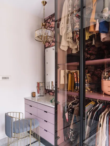 The completed pink walk-in closet, showing the vanity area next to the glass wardrobe, with a stylish chair and elegant chandelier.