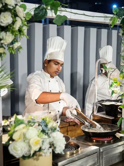 A chef at a live pasta station, expertly tossing ingredients in a pan. The surrounding floral decor shows how we integrate our cooking stations beautifully into the overall event design.