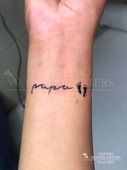 A touching "papa" tattoo in a child's handwriting style, paired with tiny baby footprints. A deeply personal and sentimental piece.