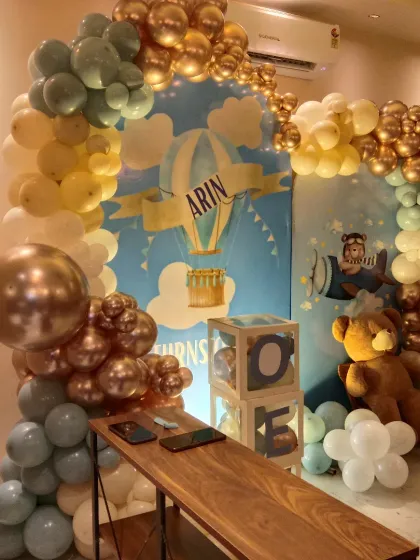 A sweet teddy bear and hot air balloon theme for a first birthday. The backdrop features a sky-blue design, surrounded by a balloon arch in pastel yellow, blue, and metallic gold.