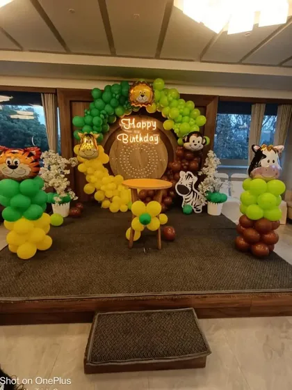 A stage setup with a jungle theme, featuring a balloon arch, animal foil balloons, and a "Happy Birthday" neon sign.