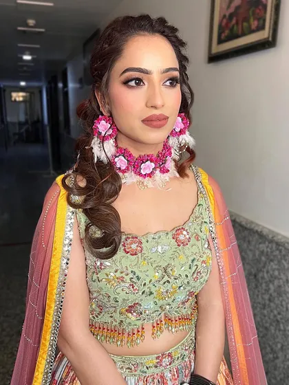 A dreamy Mehendi look. The client is wearing a floral pastel lehenga with colorful jewelry and curled hair. The makeup is very minimal, just enhancing her beauty.