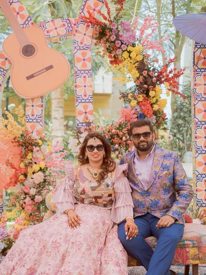 The couple at their 'Mexican Fiesta' Mehendi, seated against a backdrop of colorful flowers and a guitar prop.