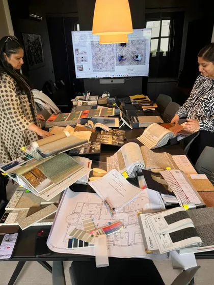 A view of the design studio during the material selection phase. The table is covered with fabric swatches, flooring samples, and architectural plans, showing the detailed and collaborative nature of the process.