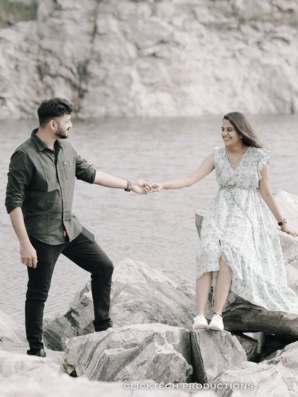A couple holds hands while standing on rocks by a lake, creating a romantic and scenic pre-wedding portrait.