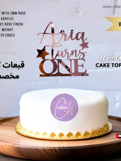 A mockup of the "Aria turns ONE" topper in rose gold on a simple white cake, showcasing its elegant design.