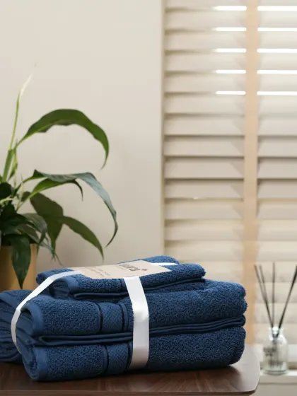 A set of navy blue towels in a softly lit bathroom setting. The warm light and background plant create a spa-like, relaxing atmosphere.