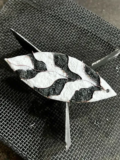 An enamel piece during the firing process, resting on a mesh before going into the kiln where the glass powder melts and fuses to the copper.