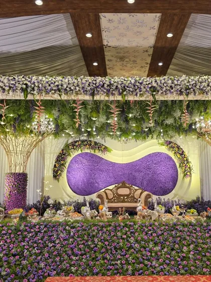A stunning stage design dominated by a sea of purple flowers. The backdrop features a unique, modern curved element covered in purple blooms, flanked by artistic tree-like structures and a foreground filled with purple flowers.