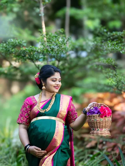 A serene outdoor maternity portrait of an expecting mother in a beautiful green and pink saree, holding a basket of flowers in a lush garden.