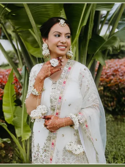 A smiling bride in a white and gold outfit adorned with floral jewelry stands against a lush green background, looking fresh and radiant.
