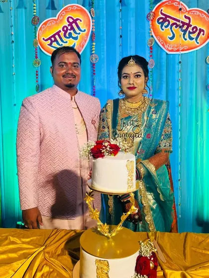 A happy couple posing with their two-tier engagement cake. The cake features a unique stand and is decorated with fresh red roses and gold leaf details, perfectly matching their festive attire.