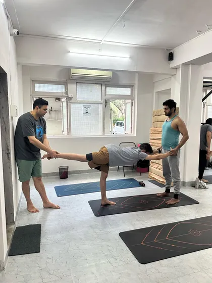 An instructor and a partner help a student find his alignment in Virabhadrasana III, using the wall for added stability.