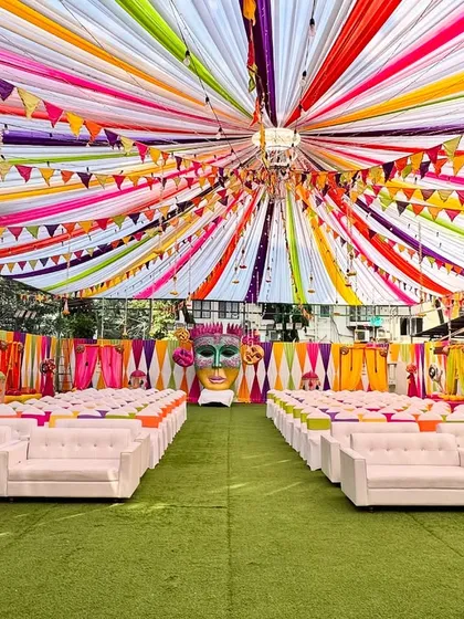 A full view of a Mehendi celebration with a ceiling of radiating multicolor drapes and festive flags. The open layout with clean white sofas creates a fun and energetic vibe.