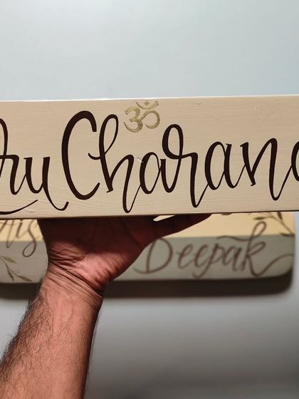 Holding up a freshly painted name plaque, "Guru Charana." The clean calligraphy and subtle Om symbol were designed to match the client's calm and spiritual vibe.