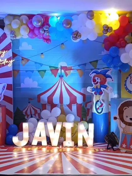 A vibrant circus carnival theme for Javin's birthday. The setup features a classic red-and-white striped circus tent backdrop, character cutouts of clowns and animals, and bright marquee letters.