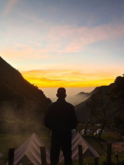 A silhouette of a person watching the sunrise over the tents and clouds at the Munnar camp.