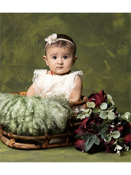 A 6-month-old baby, sitting up in a basket against a rich green backdrop. The deep red flowers add a beautiful pop of color.