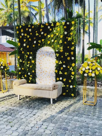 For this outdoor haldi ceremony, I created a vibrant photo spot with a lush greenery wall dotted with yellow and white flowers. The design includes a comfortable seating area and matching floral stands, all set against a beautiful natural backdrop of palm trees.