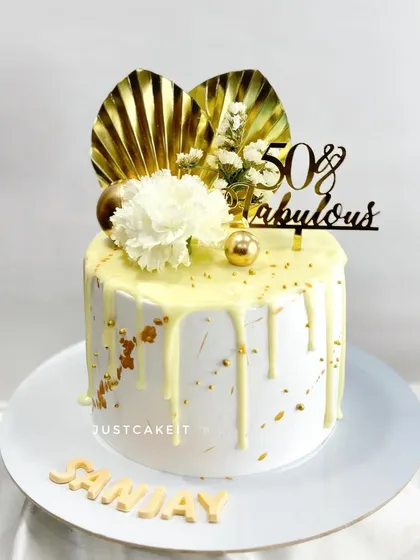 A "50 & Fabulous" birthday cake. This design features a white chocolate drip, gold leaf accents, and a modern topper with a white carnation.