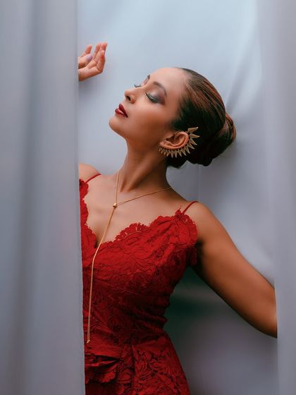 An artistic and glamorous shot featuring a model in a red lace dress. The use of white curtains for framing creates a sense of mystery and highlights the bold color and jewelry.
