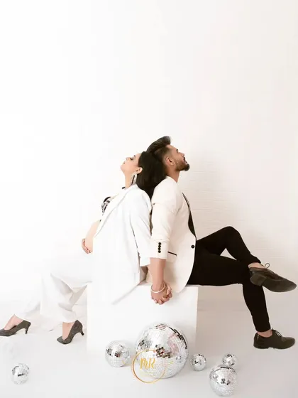 An artistic, high-fashion pose. The couple sits back-to-back in contrasting black and white outfits, creating a strong, symmetrical, and modern maternity photograph.