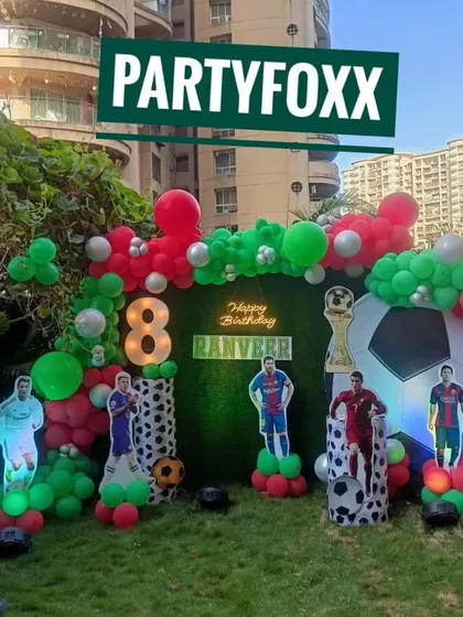 An outdoor football party setup for Ranveer's 8th birthday, with a green hedge backdrop, player cutouts, and a red and green balloon arch.