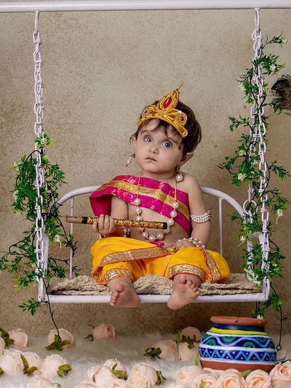 A full view of the swing setup for a Janmashtami shoot, with the little Krishna holding his flute. The floor is decorated with flowers, adding to the festive feel.