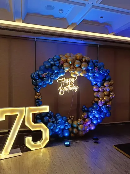 A grand 75th birthday celebration with a blue and gold theme. A full ring of metallic balloons frames a "Happy Birthday" neon sign, complemented by large light-up numbers for the milestone.
