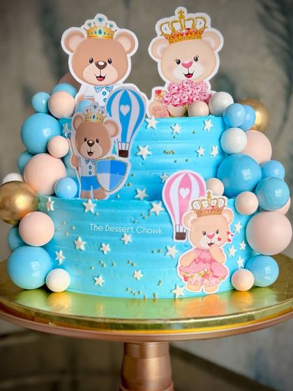 A sweet two-tier cake for a baby shower celebration. The blue buttercream is decorated with cute teddy bear and hot air balloon toppers, surrounded by a cascade of blue, white, and gold spheres.
