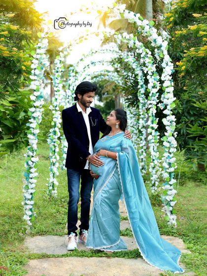 A romantic couple's portrait under a floral archway. The mother-to-be is wearing a beautiful light blue saree that drapes elegantly.