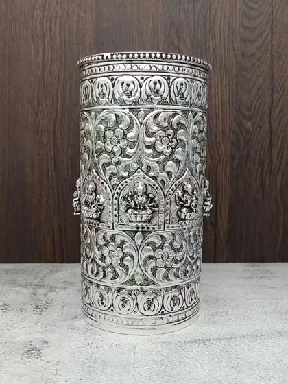 A tall, antique German silver container or glass, with intricate floral patterns and carvings of Goddess Lakshmi.