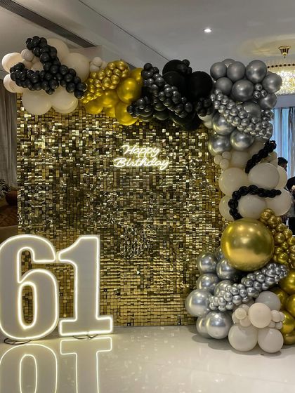 A luxurious decoration for a 61st birthday, featuring a gold sequin wall. The decor is enhanced by a black, gold, and silver balloon garland and large, brightly lit marquee numbers.