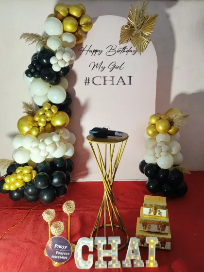 I can create personalized backdrops like this modern arch panel for a birthday celebration. The setup is framed with a black, white, and gold balloon garland and features the name 'CHAI' in light-up letters for a fun, custom touch.